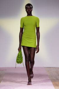 Model walks a runway in a lime green, short-sleeve bodycon dress, holding a matching ruffled green bag.