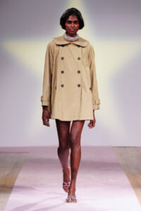 Model on a runway wearing a beige double-breasted trench coat with a wide collar, pearl choker, and white strappy sandals.