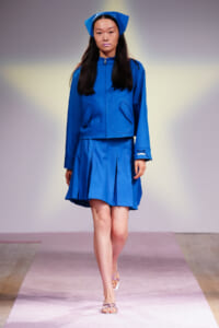 Runway model wearing a bright blue zip-front jacket with large pocket flaps and a matching pleated skirt, plus a blue headscarf.