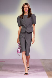 Model walking a fashion runway in a gray tailored suit with puff sleeves, carrying a shiny pink handbag and wearing black sandals.