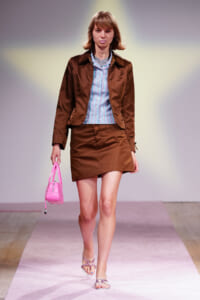Model walking a fashion runway in a brown matching blazer and mini skirt with a light blue striped shirt and pink handbag.