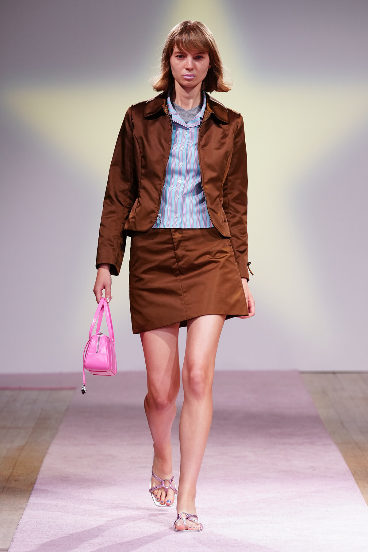 Model walking a fashion runway in a brown matching blazer and mini skirt with a light blue striped shirt and pink handbag.