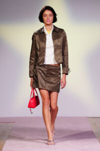 Female model on a runway wearing a brown satin cropped blazer and matching mini skirt over a white button-down shirt, carrying a bright red handbag.