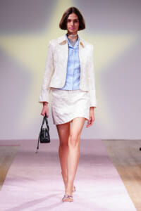 Model walking a fashion runway in a cream textured blazer and matching mini skirt, paired with a light blue gingham shirt and a small black handbag.