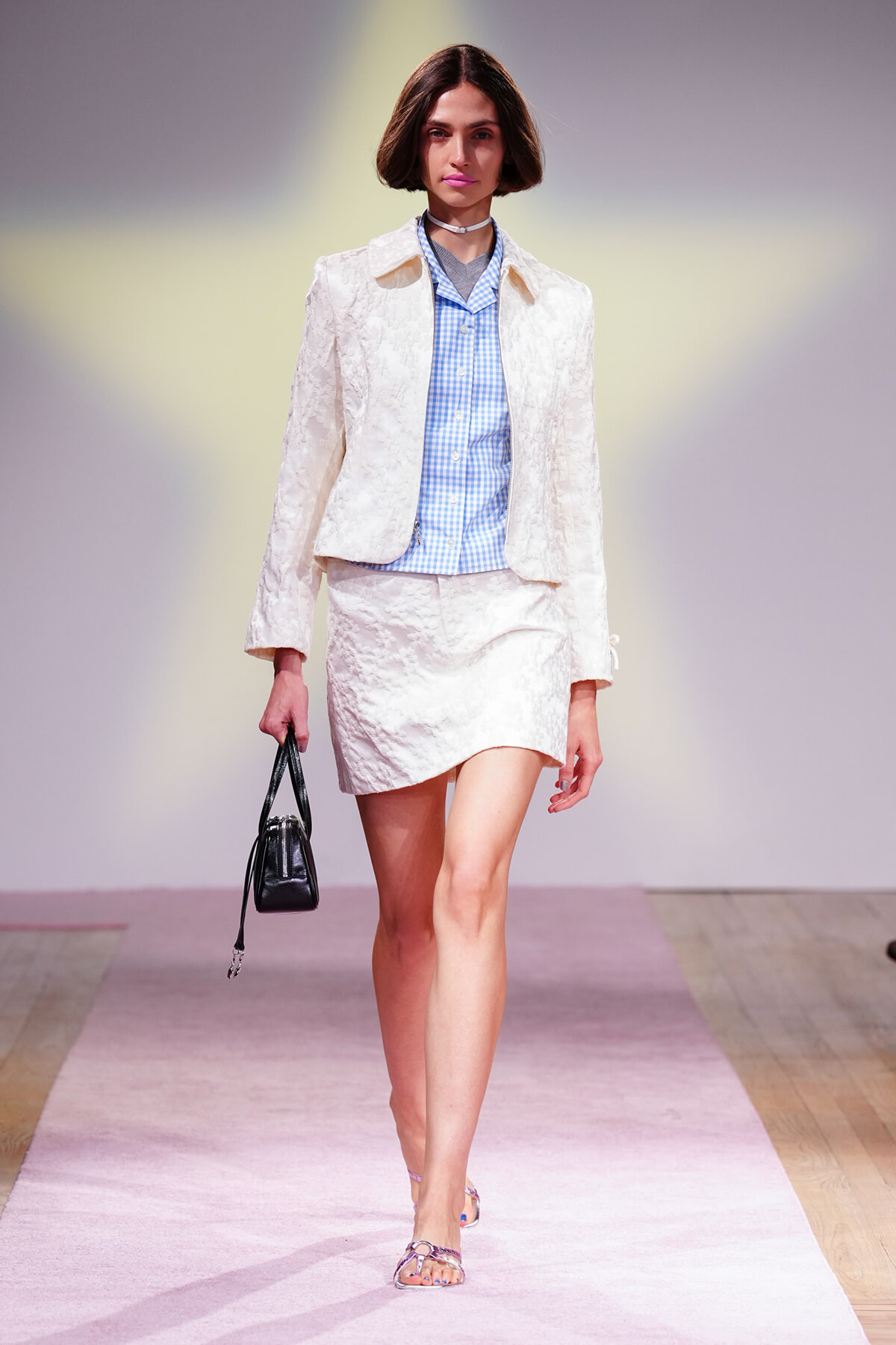 Model walking a fashion runway in a cream textured blazer and matching mini skirt, paired with a light blue gingham shirt and a small black handbag.