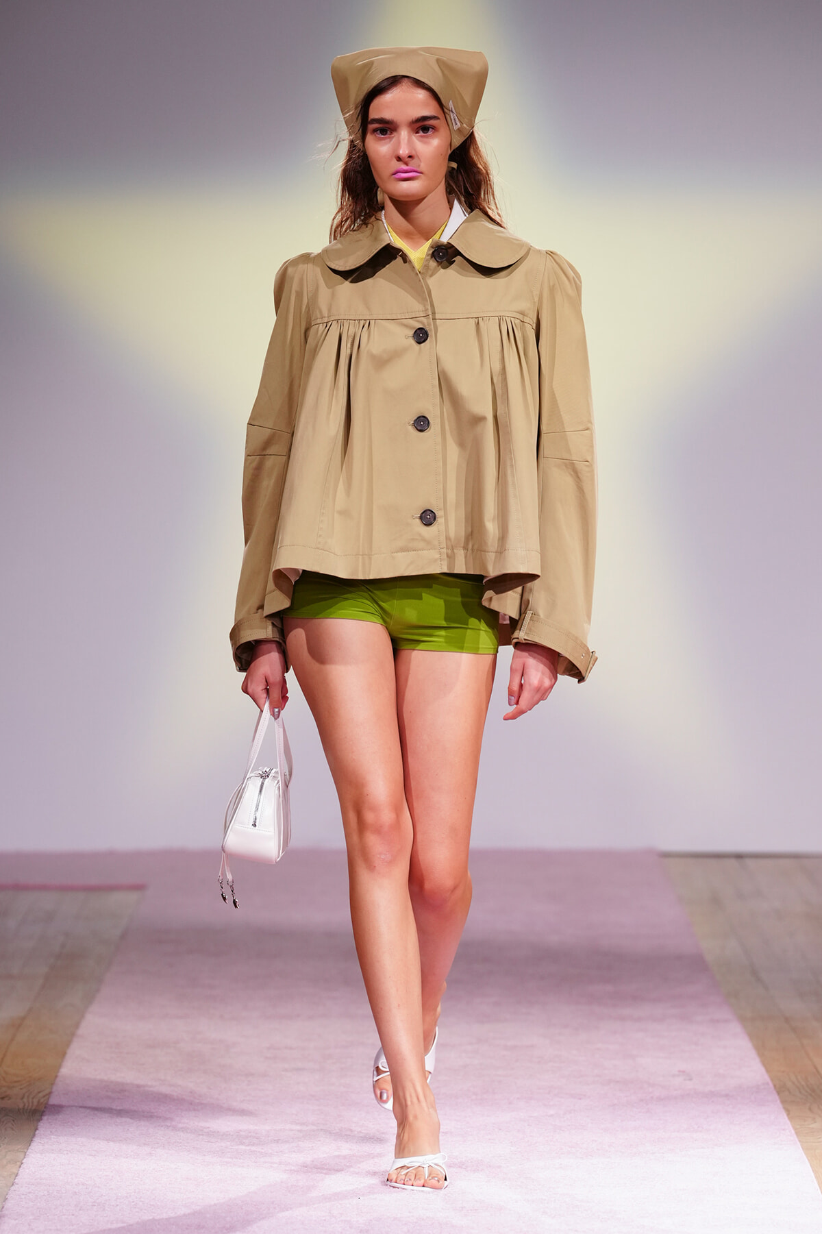 Model walking a runway in a beige roomy trench-style jacket, green shorts, white sandals, and a tan hat, carrying a small white bag.