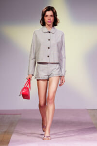 Model walking a fashion runway in a light pastel checkered suit with a cropped jacket and matching shorts, carrying a bright pink handbag and wearing pink sandals.