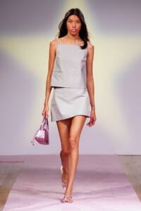 Model walks a runway in a pastel blue-and-white gingham two-piece set (top and mini skirt), carrying a pink metallic handbag.