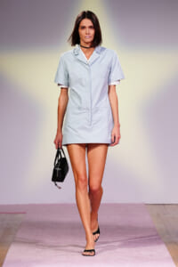 Female model walking the runway in a light blue short-sleeve shirt dress, carrying a small black handbag and wearing black flat sandals.