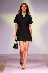 Female model walks the runway in a black short-sleeve dress over a blue gingham shirt, accessorized with a black choker and a small black handbag.