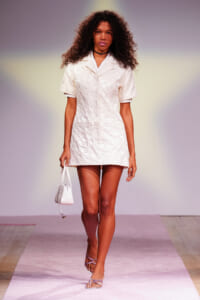 Model walking a fashion runway in a short, textured white dress, carrying a small white handbag and wearing lilac sandals on a pink runway.