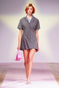 Model walking a runway in a gray short-sleeve dress, carrying a pink handbag and wearing strappy sandals.