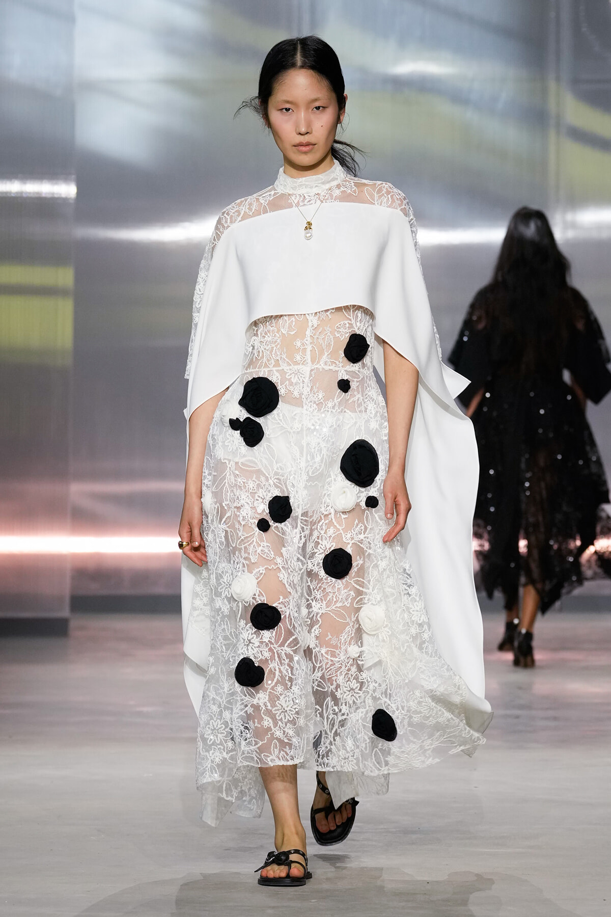 Model walks the runway in a white lace dress with sheer panels, floral appliques, and a cape诚信.