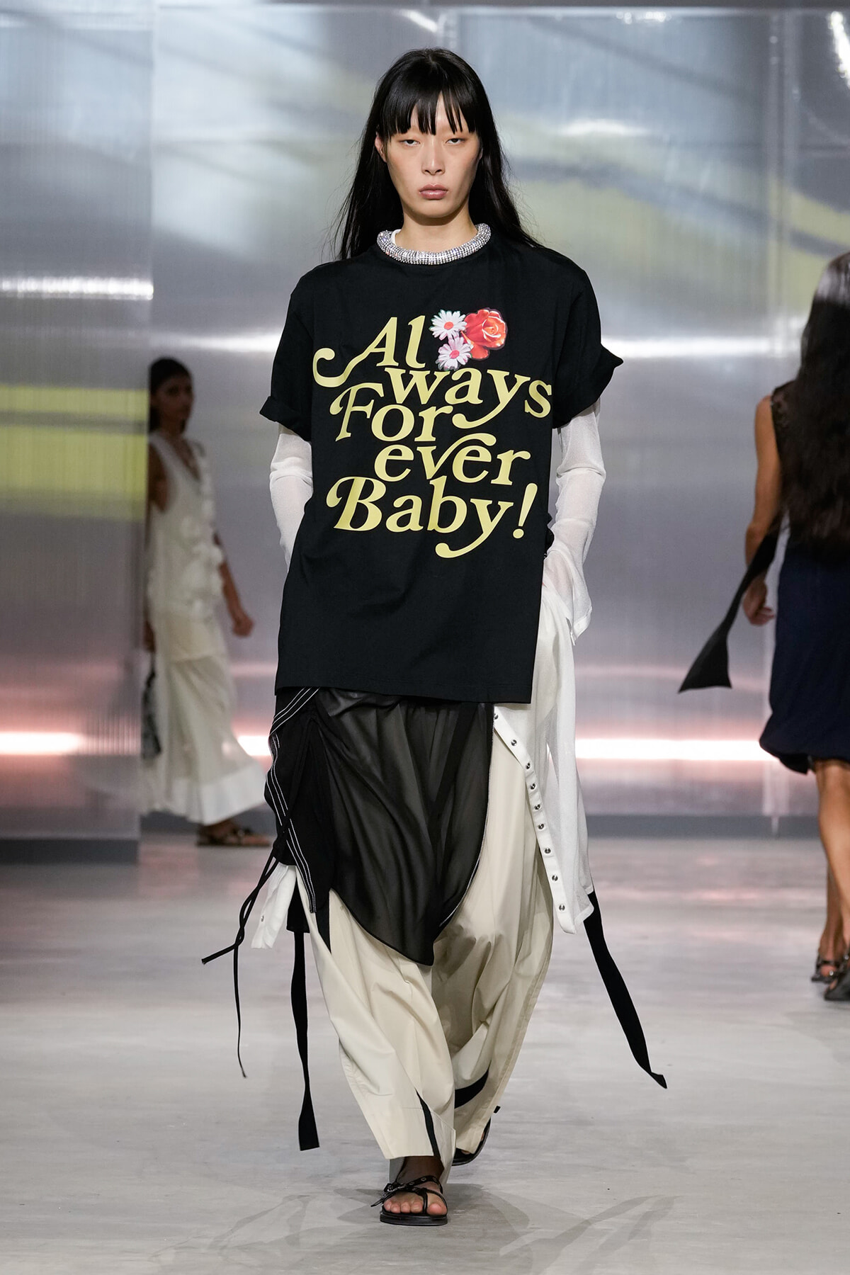 Model on a runway wearing a black graphic T‑shirt with gold lettering and flower accents, paired with layered white sleeves and draped beige/black pants.