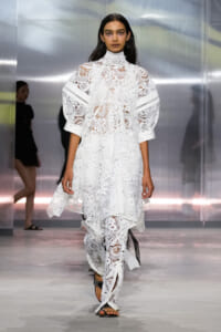 Female model walking down a runway in a white lace high-neck dress with voluminous sleeves and a layered, sheer skirt.