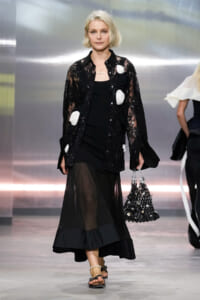 Model walking a runway in a black lace coat with white fabric rosettes over a black dress, holding a beaded black handbag.