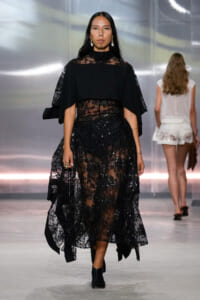 Model walking the runway in a black lace evening gown with a sheer floral overlay and cape-like sleeves.