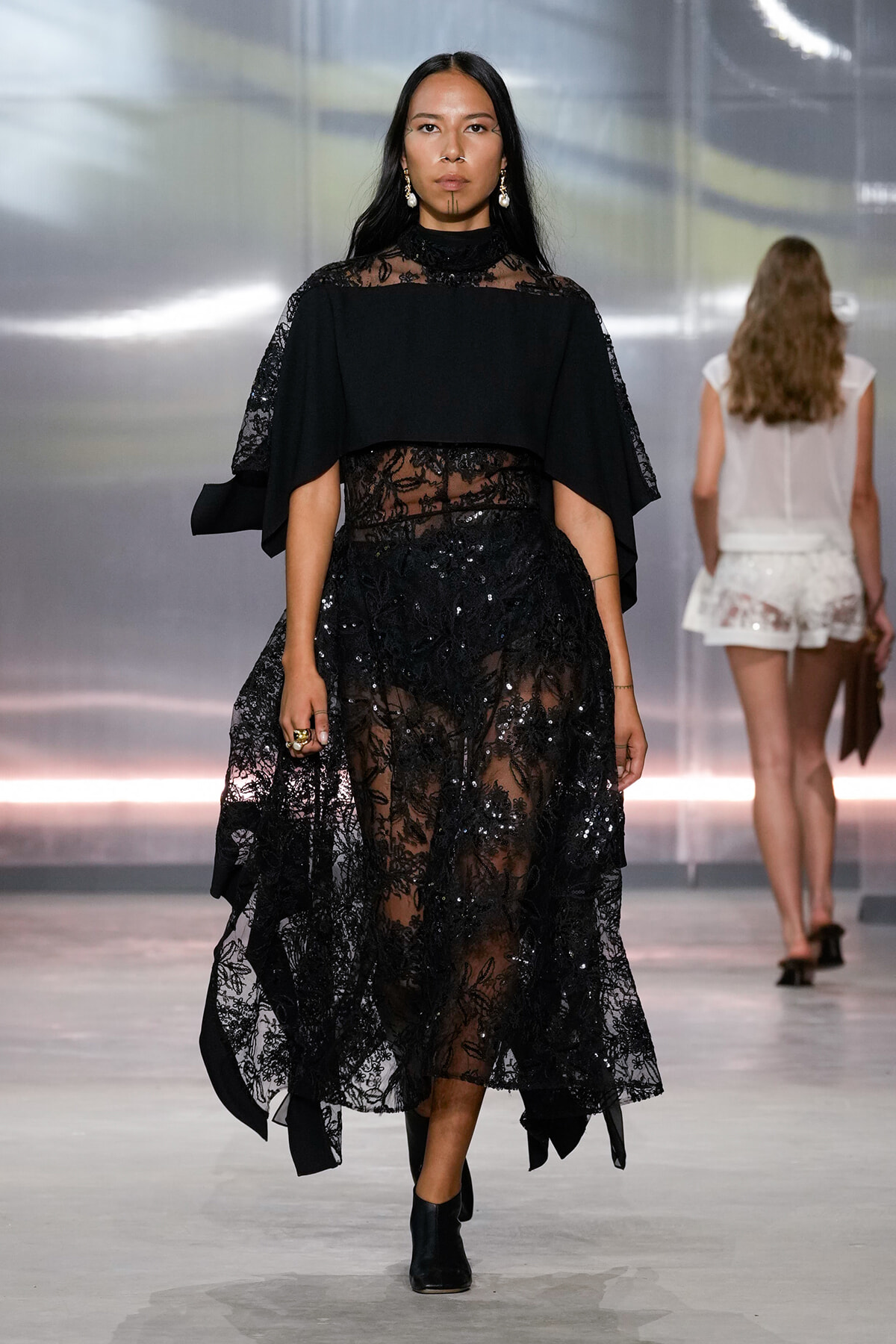Model walking the runway in a black lace evening gown with a sheer floral overlay and cape-like sleeves.