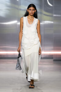 Model on a runway wearing a sleeveless white dress with 3D floral rosettes, carrying a silver beaded handbag and wearing black sandals