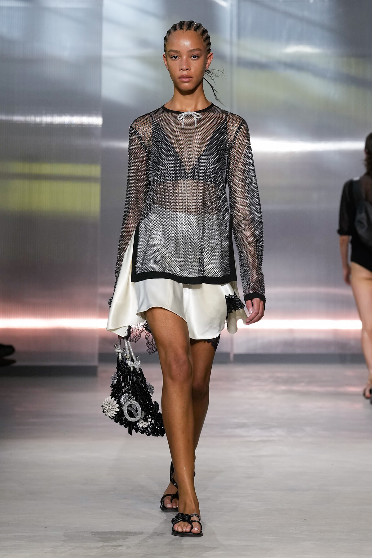 Model walks a runway in a sheer black mesh top over a black bra, paired with an asymmetric white skirt and black embellished handbag.