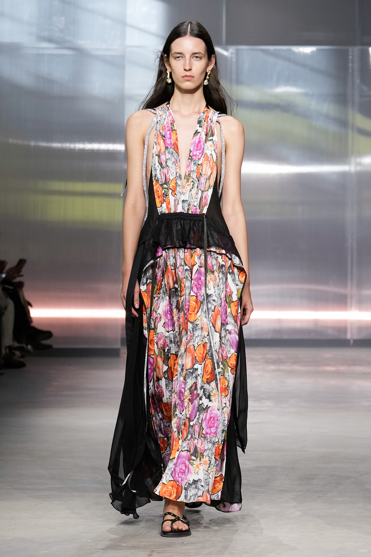 Model walking a floral maxi dress with sheer black overlays on a runway, sandals included.