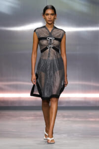 Model walking runway in a sheer mesh black-and-white dress with lace-up front panels and black trim, white sandals.