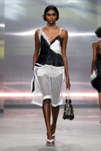 Model on a runway wearing a black-and-white satin top with lace trim and a white mesh skirt, carrying a woven net bag.