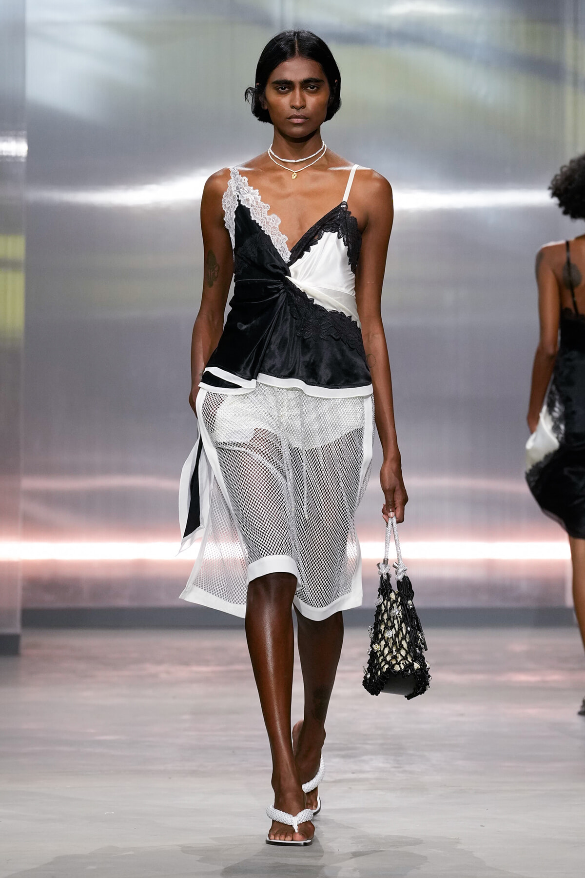 Model on a runway wearing a black-and-white satin top with lace trim and a white mesh skirt, carrying a woven net bag.