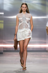 Model on a runway wearing a white mesh sleeveless top with gathered front over lace shorts, paired with brown sandals and a brown clutch.