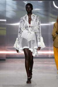 Runway model wearing a sheer white lace blouse and layered mesh skirt, walking down a metallic-backed catwalk.