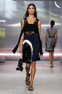 Female model walks a fashion runway in a black lace sleeveless top and a navy color-block skirt with white and black lace trim, holding a beaded black bag and wearing flat sandals.