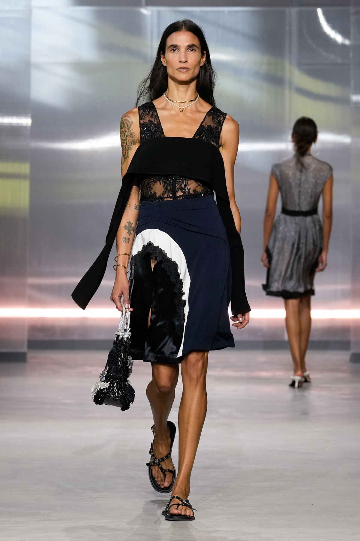 Female model walks a fashion runway in a black lace sleeveless top and a navy color-block skirt with white and black lace trim, holding a beaded black bag and wearing flat sandals.