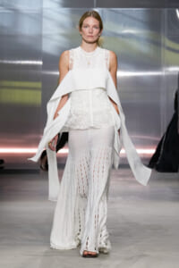 Model walking a runway in an all-white ensemble: lace sleeveless top with a large, structured cape drape over the shoulders and long, perforated wide-leg pants with slits.