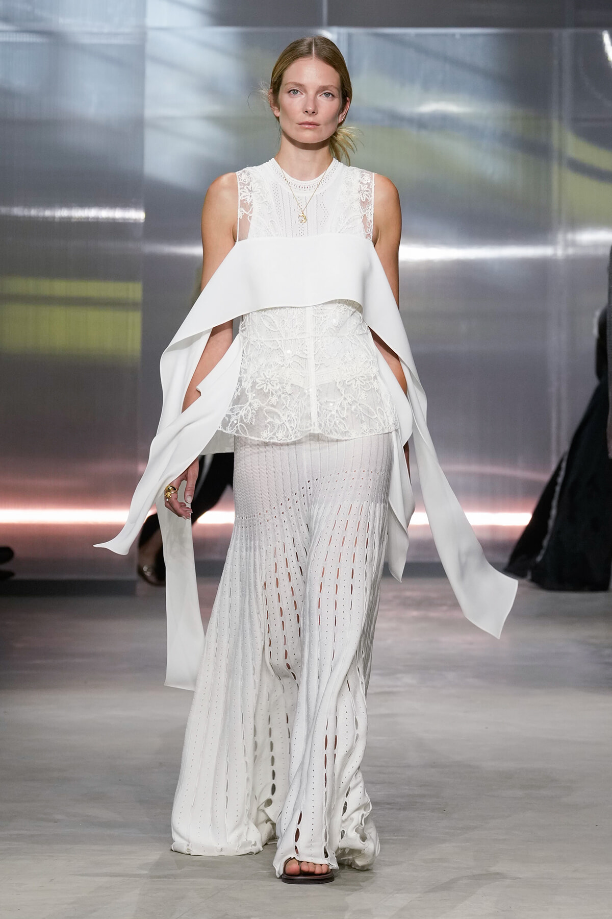 Model walking a runway in an all-white ensemble: lace sleeveless top with a large, structured cape drape over the shoulders and long, perforated wide-leg pants with slits.