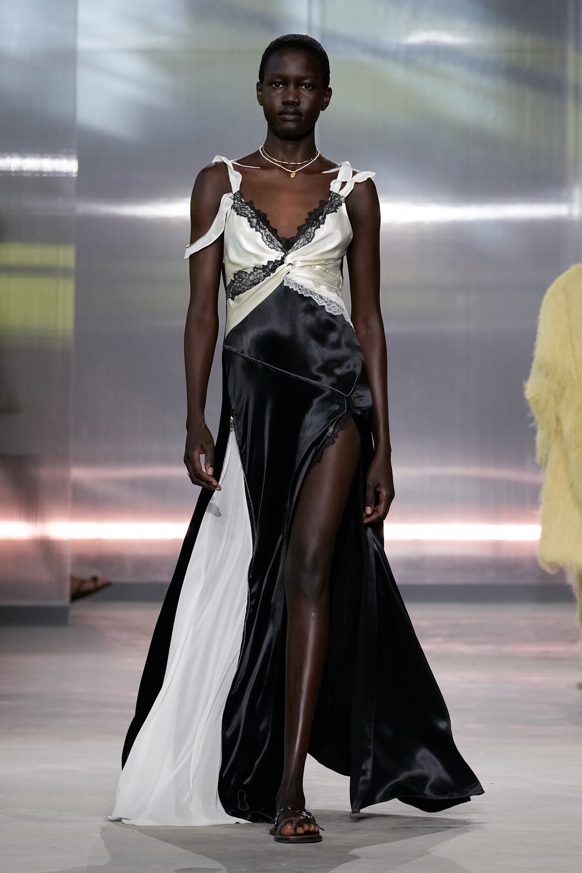 Model walking a runway in a satin black-and-white gown with lace trim and a high slit at the front.