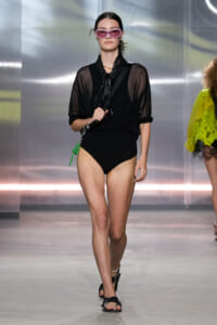Model walking the runway in a black sheer blouse over a high-cut black swimsuit, wearing pink oversized sunglasses and black sandals with a green bag charm.