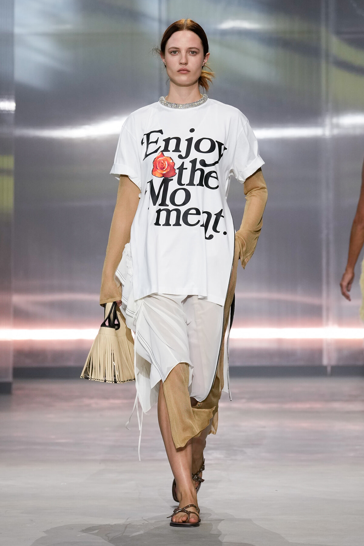 Female model walking a runway in an oversized white T-shirt with 'Enjoy the Moment' graphic and a rose, paired with beige arm warmers and a fringe handbag.