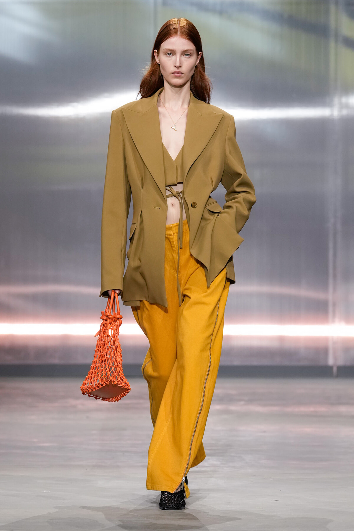 Model walking runway in oversized tan blazer, mustard wide-legged pants, carrying an orange net bag.