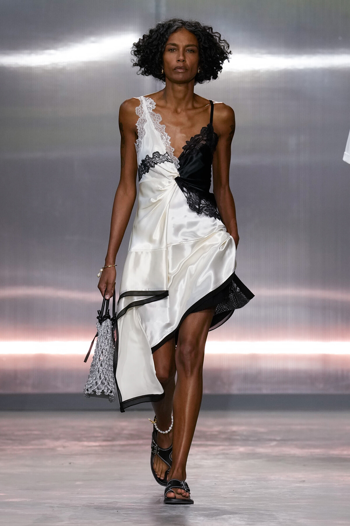 Model walks the runway in a white satin dress with black lace panels and an asymmetric hem, carrying a beaded bag.