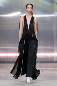 Model on a runway wearing a black, plunging V-neck gown with metallic shoulder straps and flowing sheer panels, paired with white ankle boots.
