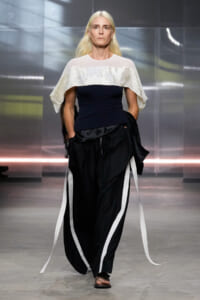 Model walks a runway wearing a navy bodice with a white capelet and voluminous black pants with white stripes, metallic backdrop behind.