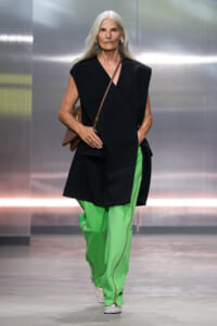 Model with long white hair walks a fashion show in a black sleeveless, asymmetrical tunic paired with bright green pants and a brown crossbody bag.