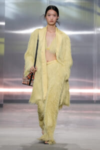 Model walks the runway in a pale yellow fringe suit with a matching blazer and crop top, carrying a patterned shoulder bag.