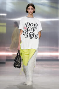 Runway model wearing a white graphic tee that says 'Don't Cry Tonight', bright neon yellow pants, shaggy white leg warmers, and a black net tote bag.