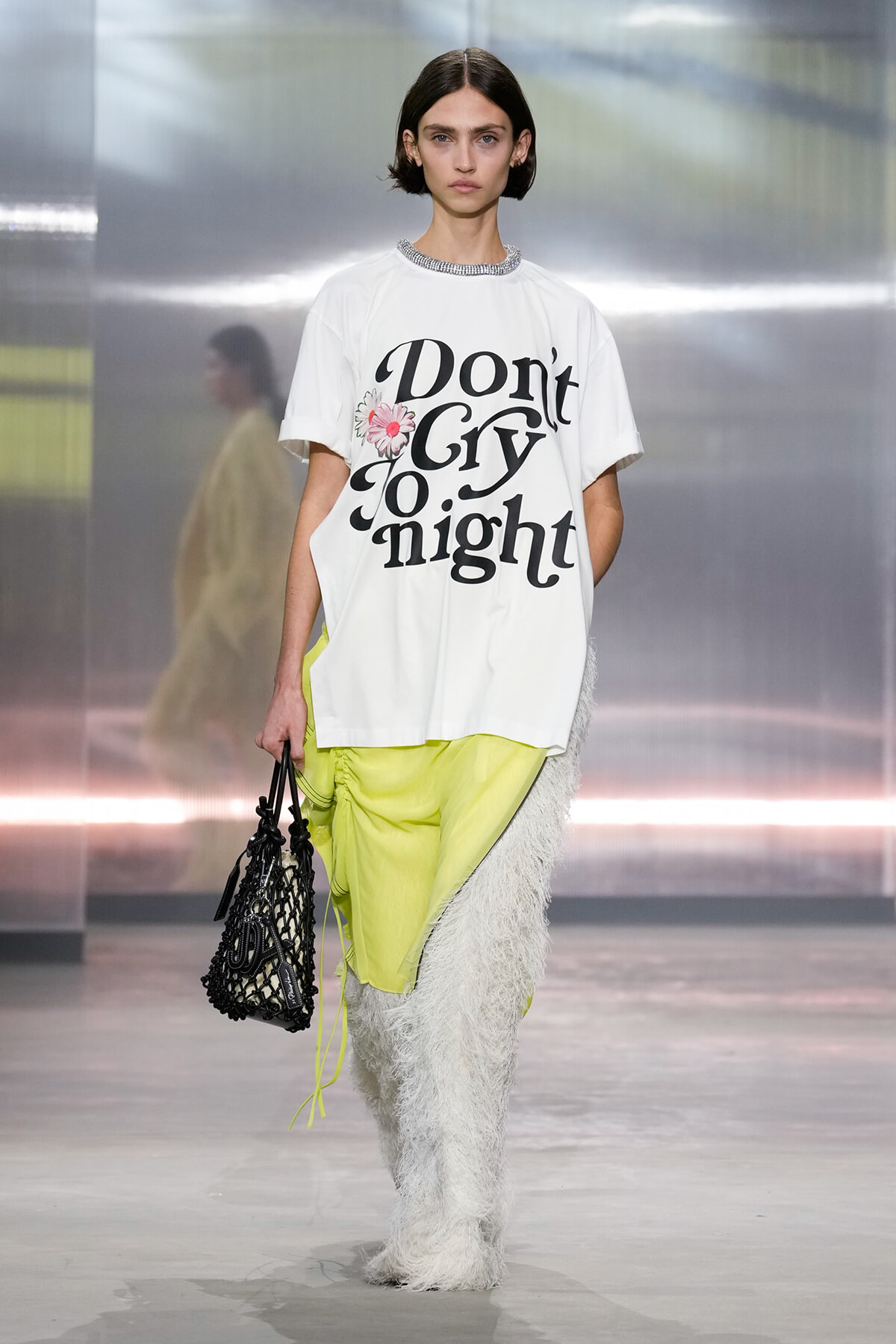 Runway model wearing a white graphic tee that says 'Don't Cry Tonight', bright neon yellow pants, shaggy white leg warmers, and a black net tote bag.