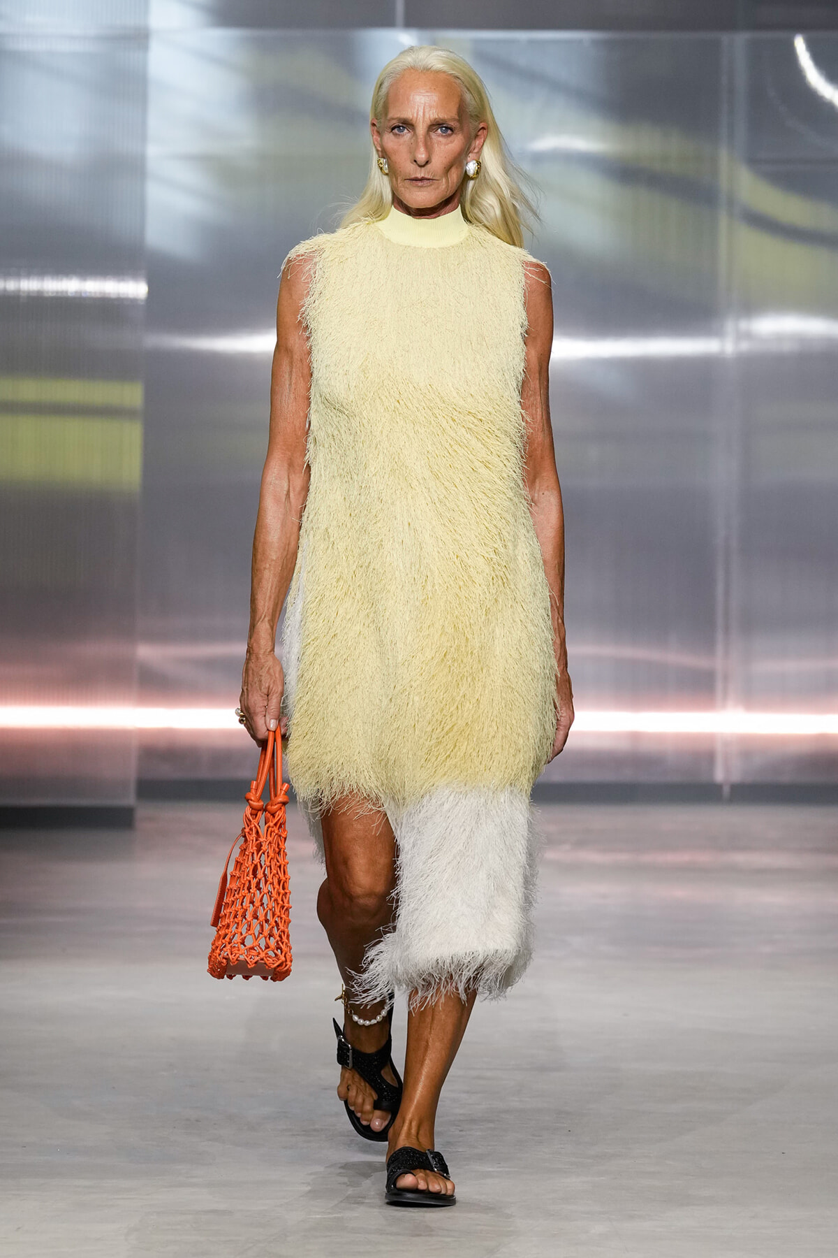 Model walking a fashion runway in a pale yellow fringe dress with a high neckline and a two-tone fringe hem, carrying an orange net bag and wearing black sandals.