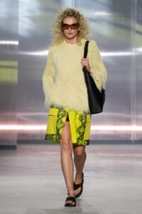 Model walking a runway in a cream fuzzy sweater and neon yellow lace-panel skirt, with a black shoulder bag and orange-tinted sunglasses.