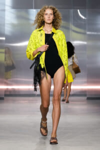 Model walking runway in a neon yellow lace cover-up over a black bodysuit, with fringe bag and brown sandals.