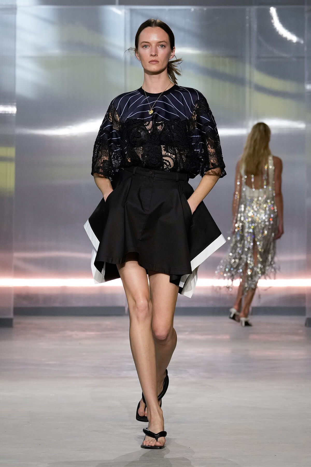 Model walks a fashion runway in a black lace blouse with striped details paired with a high-waisted, flared black skirt with white trim, neutral makeup backdrop of metallic walls and soft pink lighting.