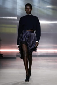 Model walking runway in a navy cropped sweater and navy striped, asymmetrical skirt with lace trim; black ankle boots and gold earrings.